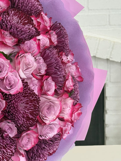 A close-up of a floral bouquet wrapped in light purple tissue paper, featuring pink and purple roses and textured chrysanthemums, set against a white brick wall background.