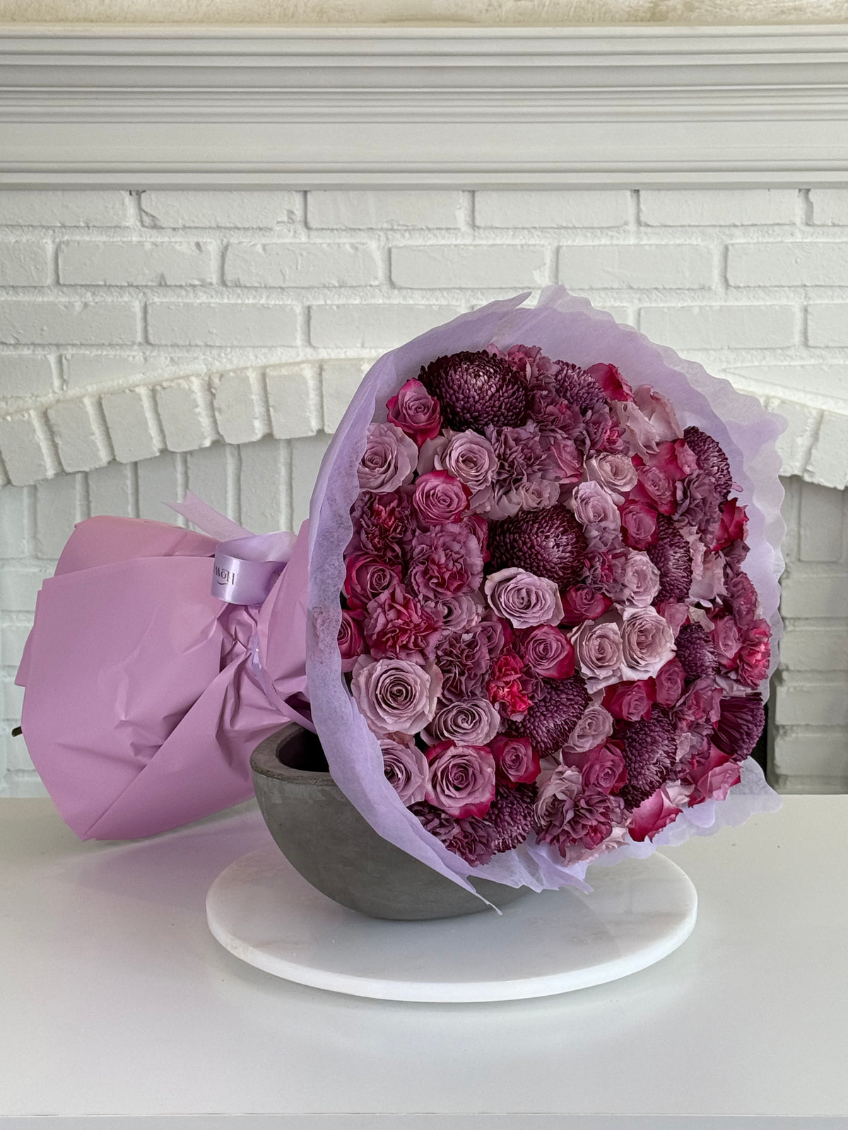 A large bouquet of pink and purple flowers wrapped in light lavender paper, displayed in a gray bowl on a white plate against a white brick fireplace background.