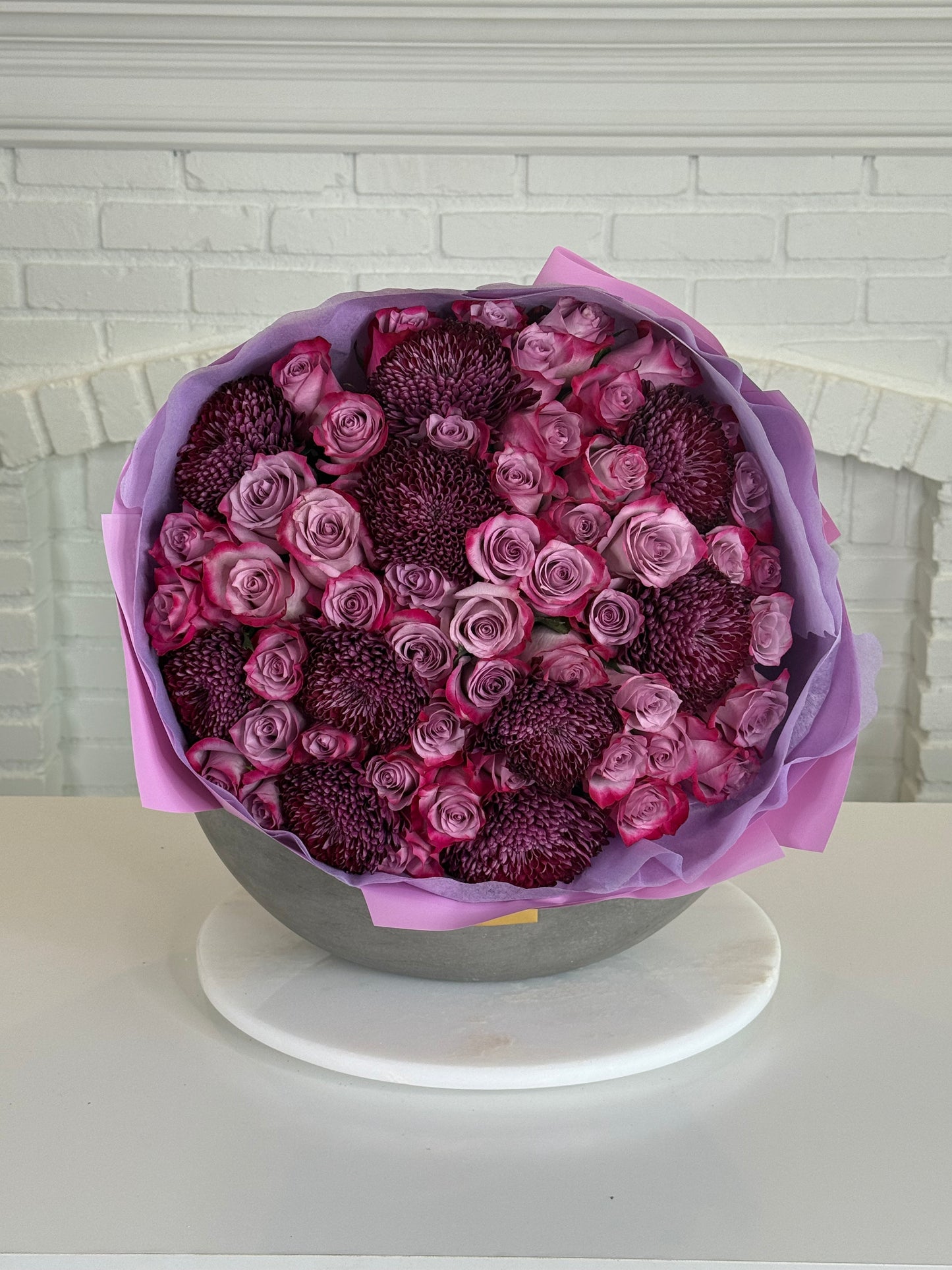 A large round floral arrangement featuring deep pink and purple roses mixed with dark purple chrysanthemums, wrapped in lavender paper and presented in a gray bowl on a white marble platter against a white brick wall background.