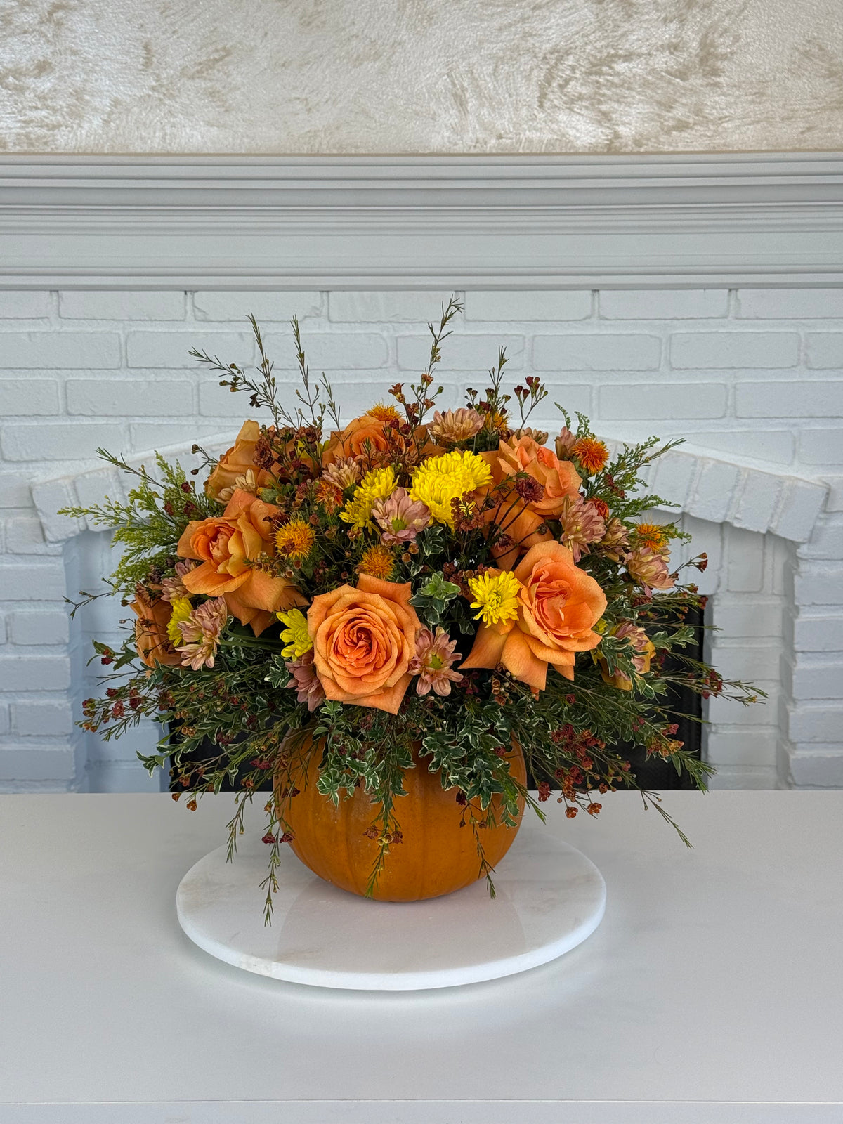 An autumn floral arrangement featuring orange roses, yellow mums, and green foliage in a pumpkin-shaped vase, displayed on a white round pedestal in front of a white brick fireplace.