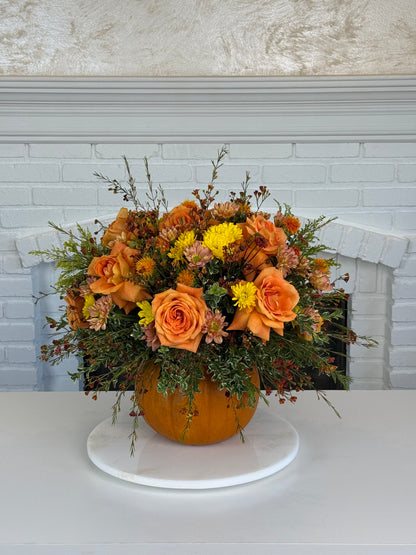 An autumn floral arrangement featuring orange roses, yellow mums, and green foliage in a pumpkin-shaped vase, displayed on a white round pedestal in front of a white brick fireplace.