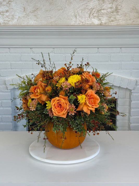 An autumn floral arrangement featuring orange roses, yellow mums, and green foliage in a pumpkin-shaped vase, displayed on a white round pedestal in front of a white brick fireplace.