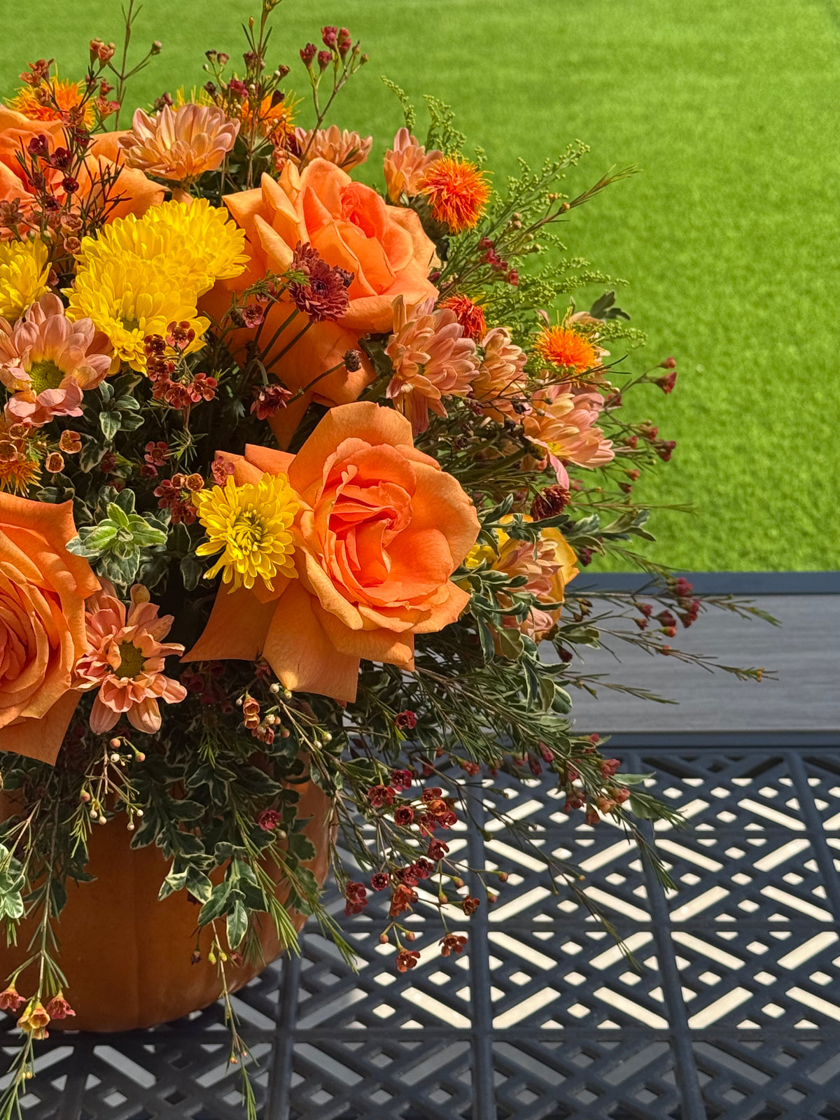 An autumnal flower arrangement in a terracotta-colored pot, featuring orange roses, yellow chrysanthemums, and green foliage, displayed on a black metal patio table with a green lawn in the background.