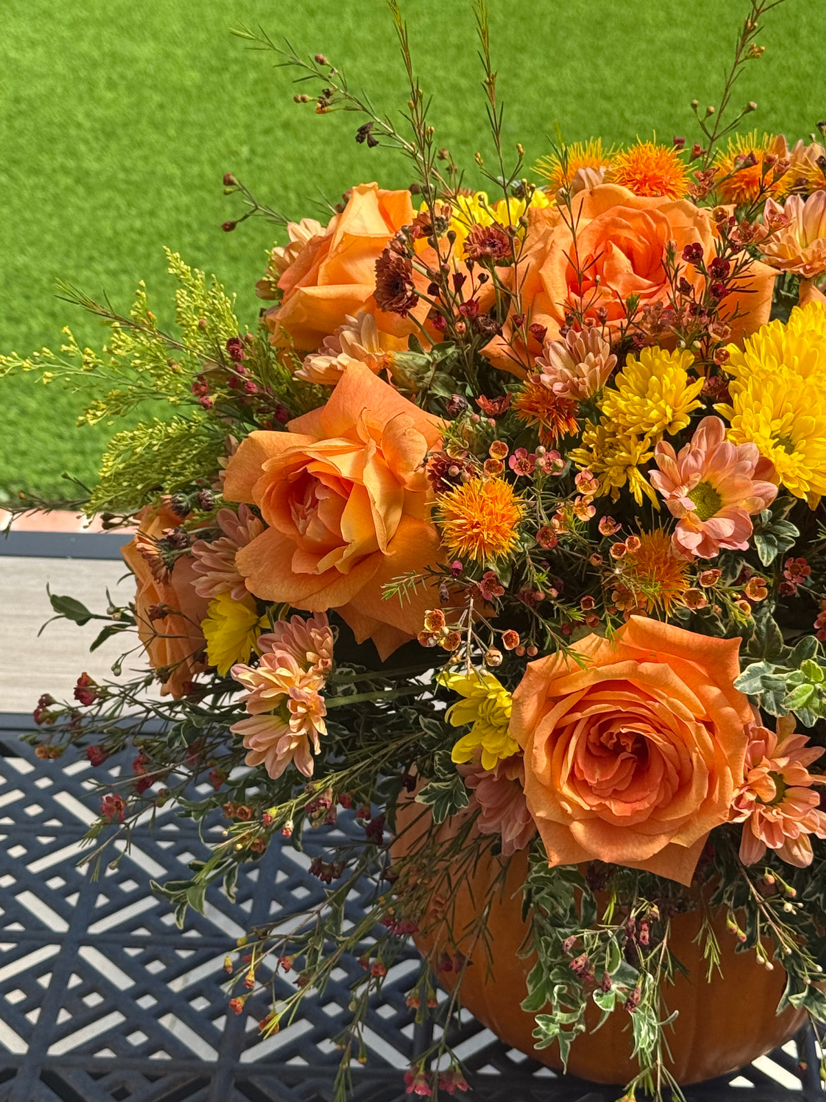 An autumnal floral arrangement in a pumpkin-shaped pot, featuring orange roses, yellow chrysanthemums, and peach daisies with green foliage, placed on a black metal patio table with a green lawn in the background.