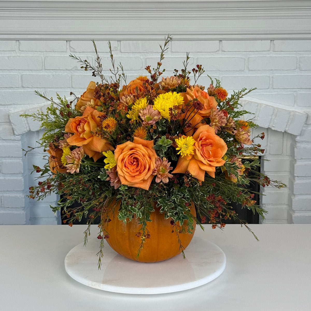 An autumn floral arrangement in a small orange pumpkin vase, set on a round white marble platter. The bouquet features orange roses, yellow chrysanthemums, and green foliage, presented against a white brick fireplace background.