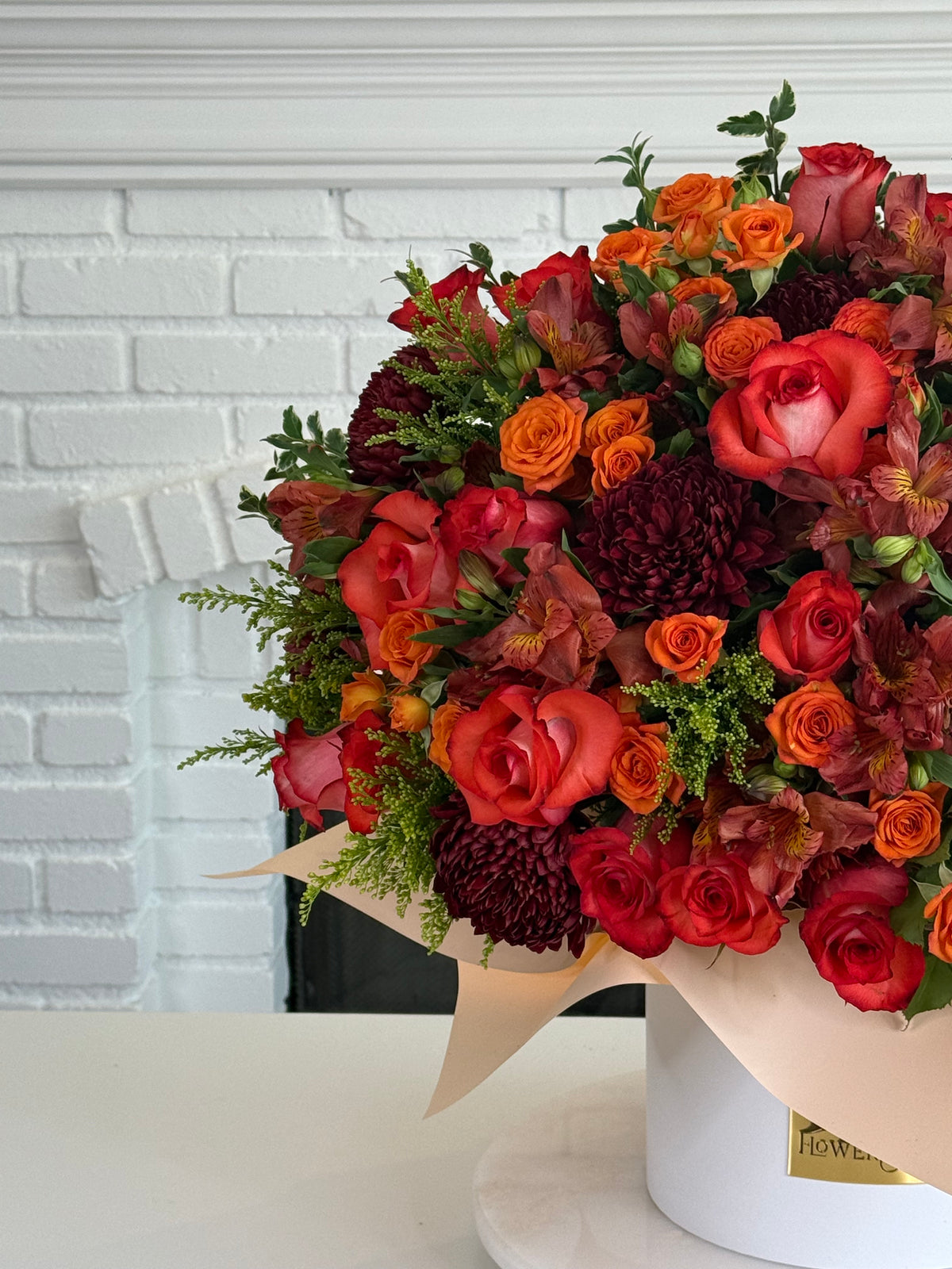 A vibrant autumnal flower bouquet in a white cylindrical pot, featuring red and orange roses, burgundy chrysanthemums, and green foliage, wrapped in beige paper against a white brick fireplace background.