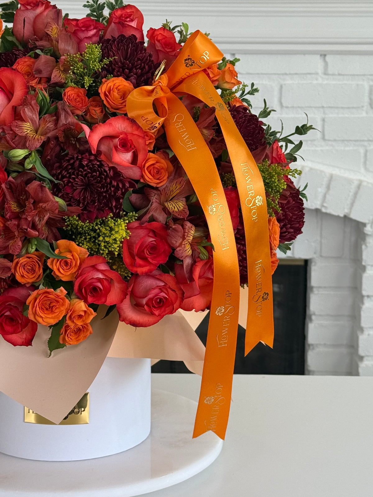 A large, vibrant floral arrangement in a white cylindrical box, featuring red and orange roses, burgundy chrysanthemums, and green foliage, tied with a prominent orange ribbon printed with the brand name 'Flowers Direct'.