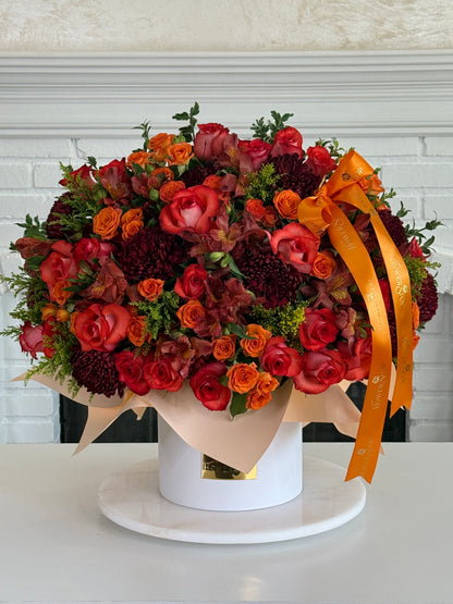 A large, vibrant floral arrangement in a round white box with a gold accent, decorated with a bright orange ribbon. The bouquet features red and orange roses, burgundy chrysanthemums, and green foliage, displayed on a white round tray against a white brick wall background.