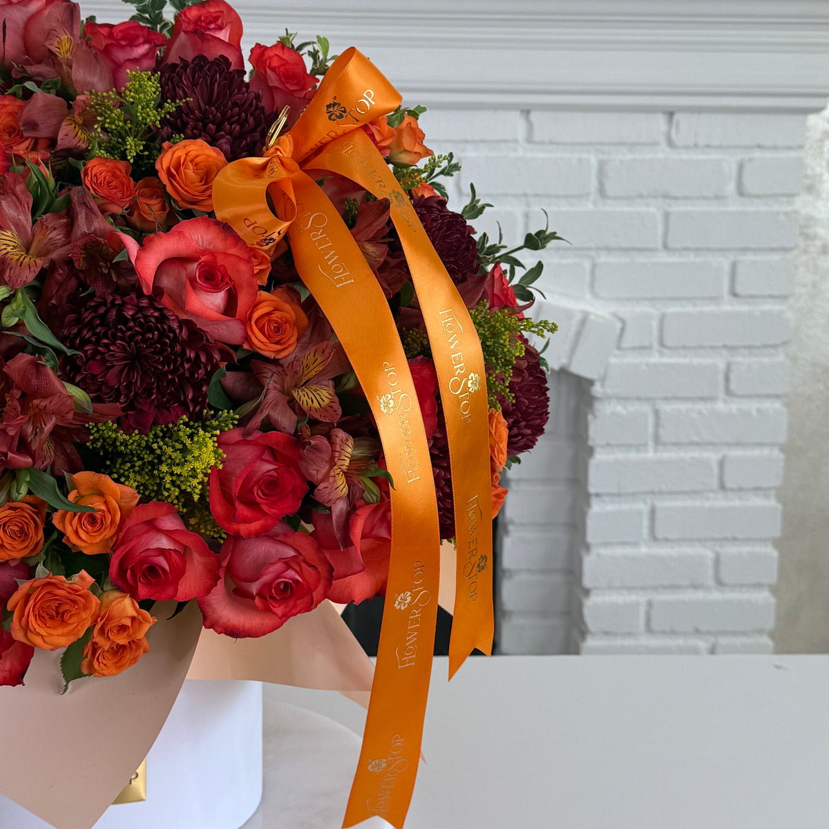 A large autumn floral arrangement in a white vase, featuring red and orange roses, burgundy chrysanthemums, and green foliage, tied with a wide orange ribbon printed with the brand name 'Southern Flower Shop'.