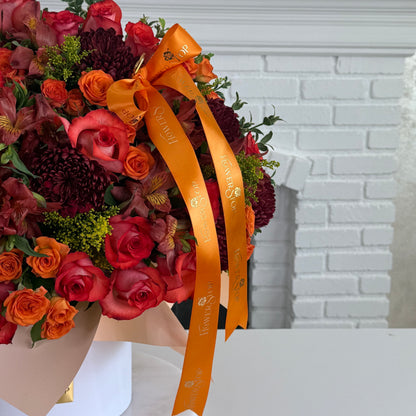 A large autumn floral arrangement in a white vase, featuring red and orange roses, burgundy chrysanthemums, and green foliage, tied with a wide orange ribbon printed with the brand name 'Southern Flower Shop'.