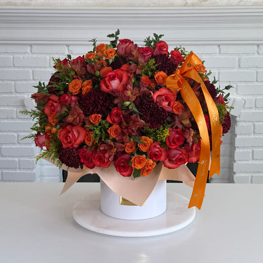 A large round floral arrangement in a white round box with a white marble base, featuring red and orange roses, burgundy chrysanthemums, green foliage, and a wide orange ribbon bow, set against a white brick wall background.