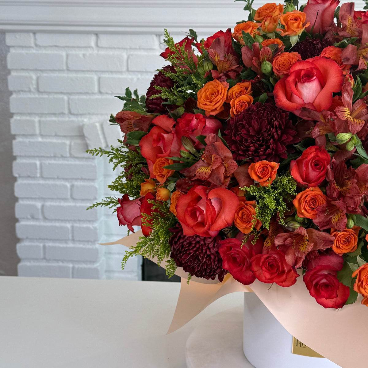 A vibrant autumn floral arrangement featuring red and orange roses, burgundy chrysanthemums, and green foliage, wrapped in a light beige paper sleeve and displayed on a white surface in front of a white brick wall.