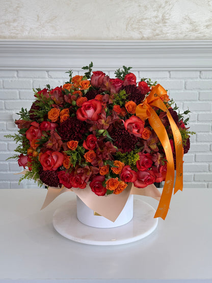A large, vibrant floral arrangement in a round white box, featuring red and orange roses, burgundy chrysanthemums, and green foliage, accented with a bright orange ribbon bow, set against a white brick wall background.