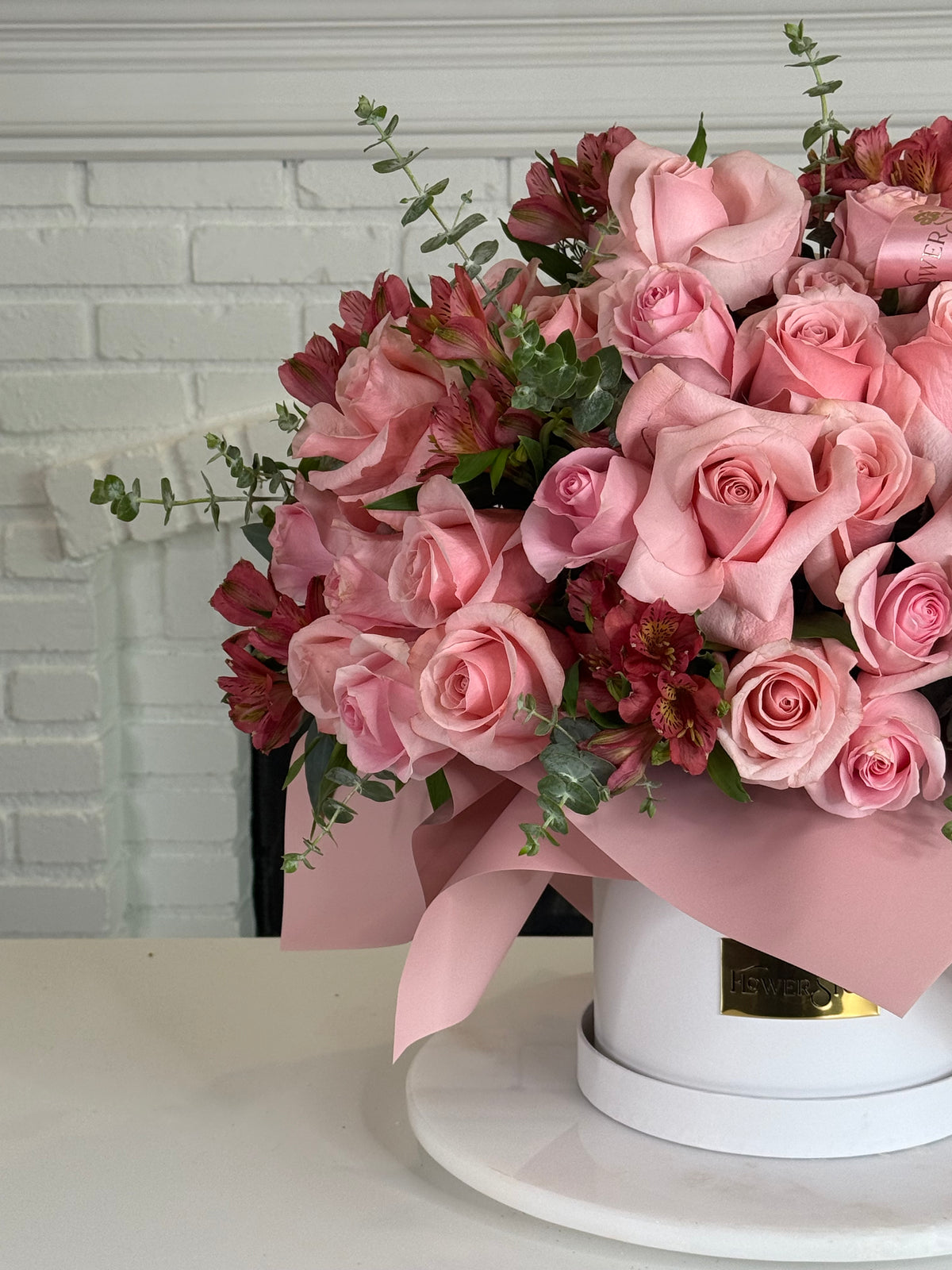 A large bouquet of pink roses and burgundy flowers wrapped in pink paper and tied with a pink ribbon, displayed in a round white box on a white surface against a white brick wall background.