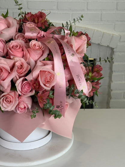 A large floral bouquet of pink roses and pink alstroemeria with green foliage, wrapped in light pink paper and tied with a pink ribbon printed with the brand name 'Flowers Direct'. The bouquet is displayed on a white round pedestal in front of a white brick fireplace.