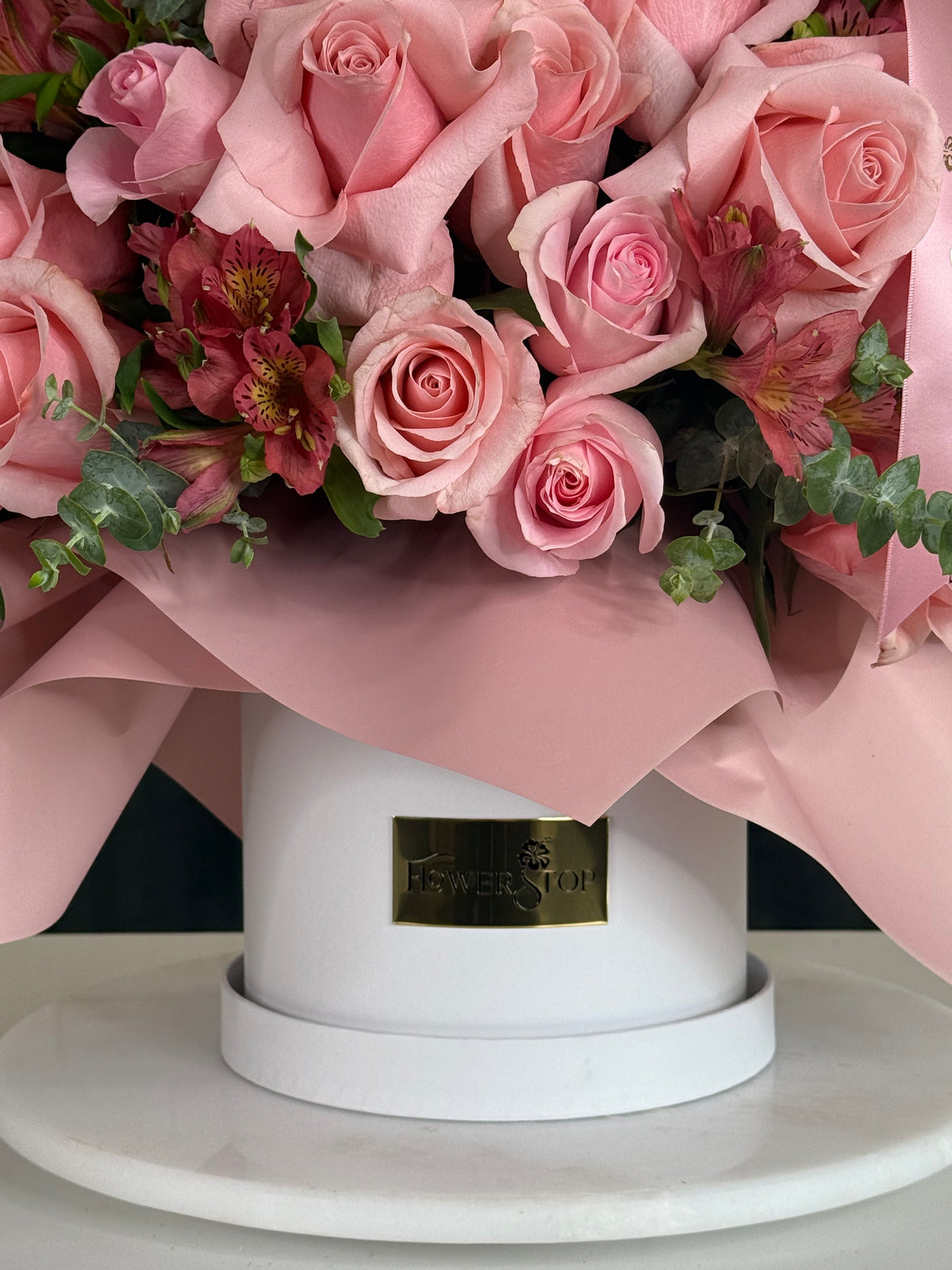 A bouquet of pink roses and pink alstroemeria flowers arranged in a round white box with pink wrapping paper and a small gold-tone Flower Shop plaque on the front, set on a white round surface.