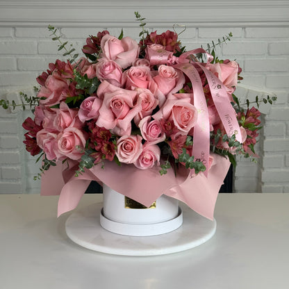 A large floral arrangement of pink roses and burgundy carnations with green foliage, tied with a pink satin ribbon, displayed in a white round box on a white marble platter against a white brick wall background.