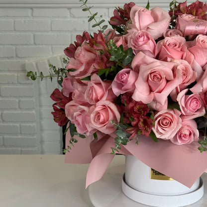 A large floral arrangement of light pink roses, dark pink alstroemeria, and green eucalyptus stems in a round white box with pink paper wrapping, set against a white brick wall background.