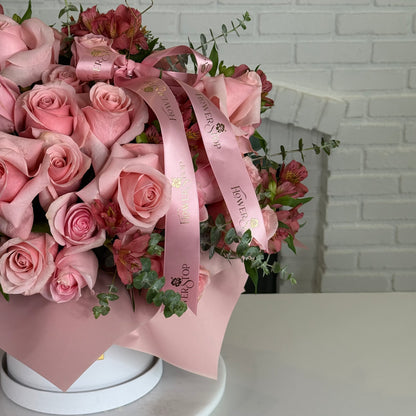 A bouquet of pink roses wrapped in light pink paper with a matching pink ribbon printed with 'Flowers Direct' and gold foil, displayed on a white surface against a white brick wall.