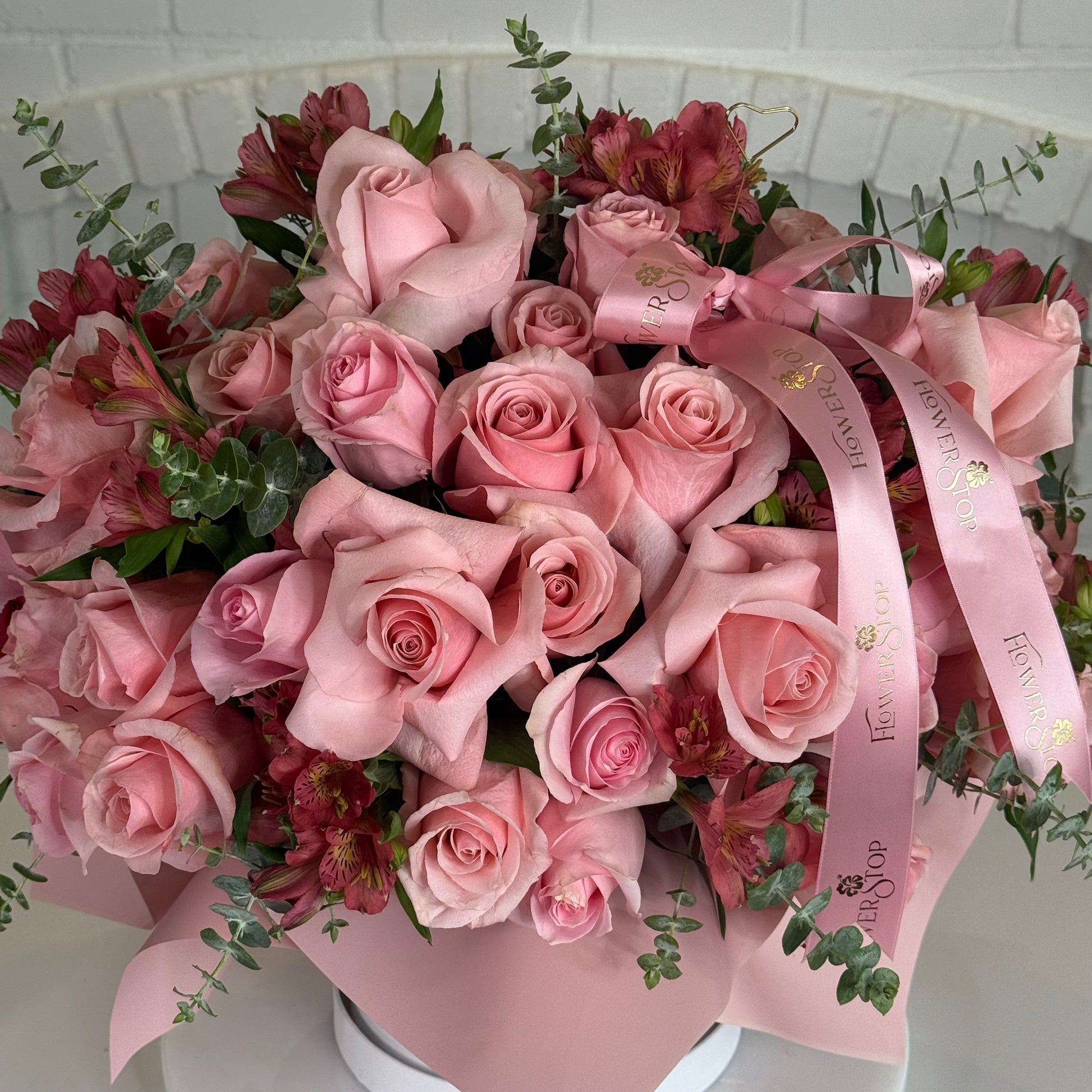 A large bouquet of pink roses and burgundy alstroemeria flowers with green eucalyptus stems, wrapped in a light pink paper sleeve with a pink ribbon printed with gold lettering, set against a white brick background.