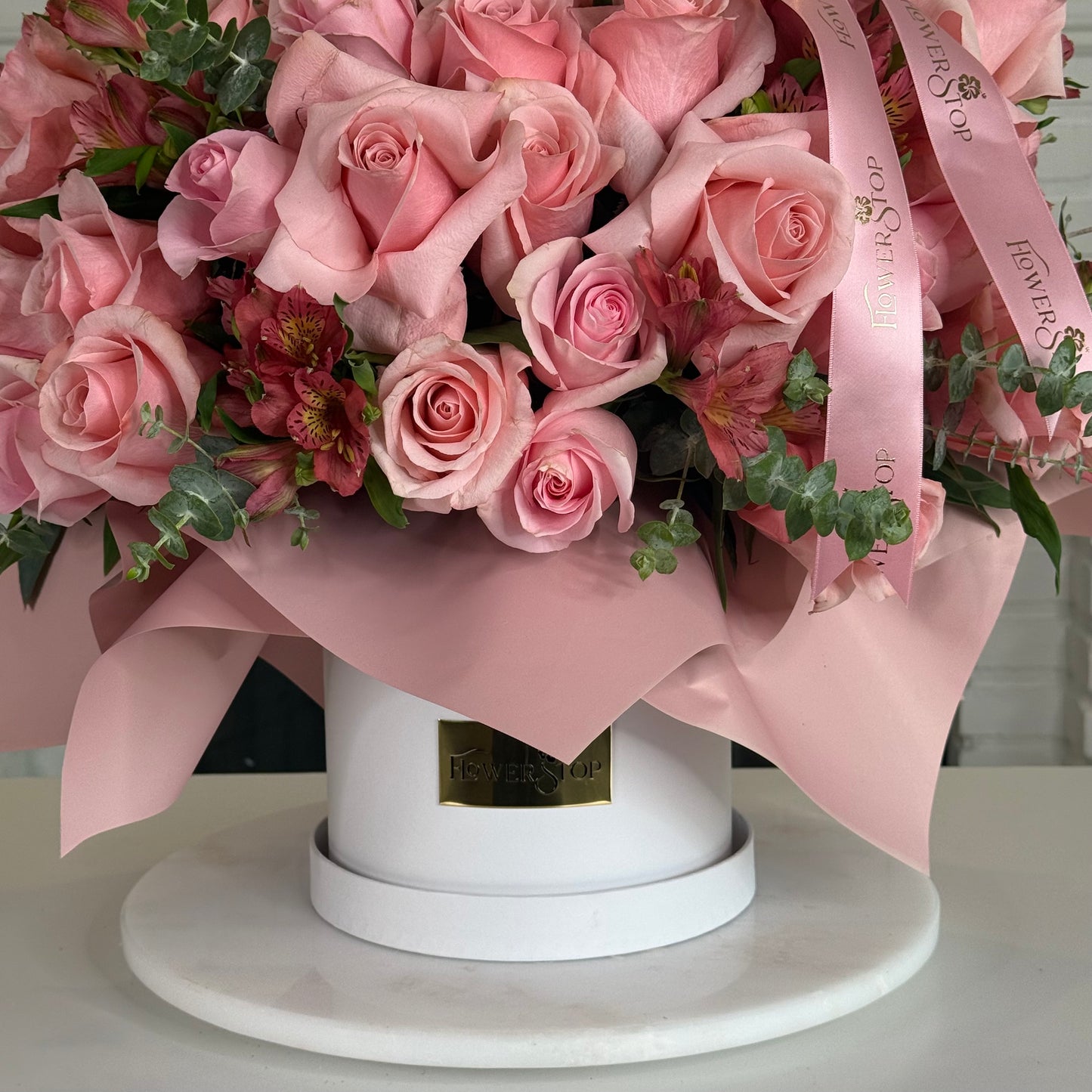 A pink floral arrangement in a round white box with pink wrapping ribbon and green foliage, photographed on a white surface against a light indoor background.