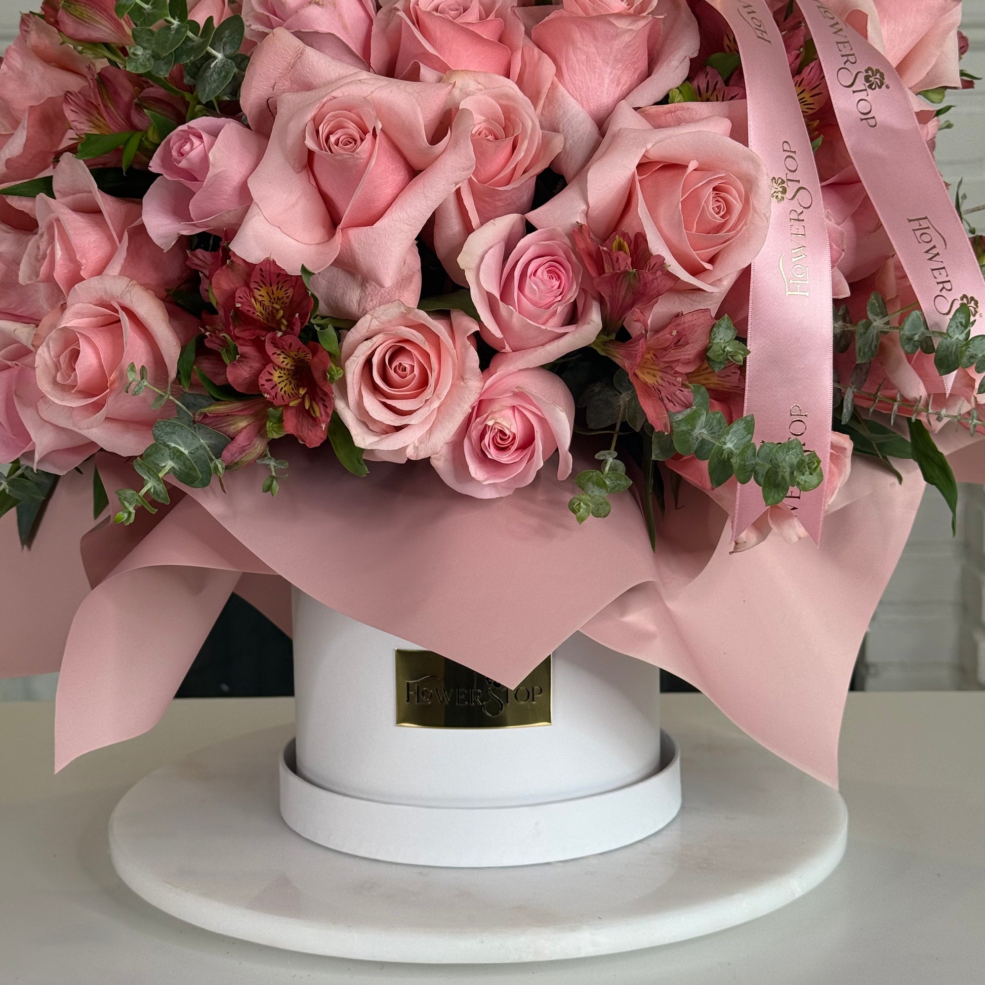 A pink floral arrangement in a round white box with pink wrapping ribbon and green foliage, photographed on a white surface against a light indoor background.
