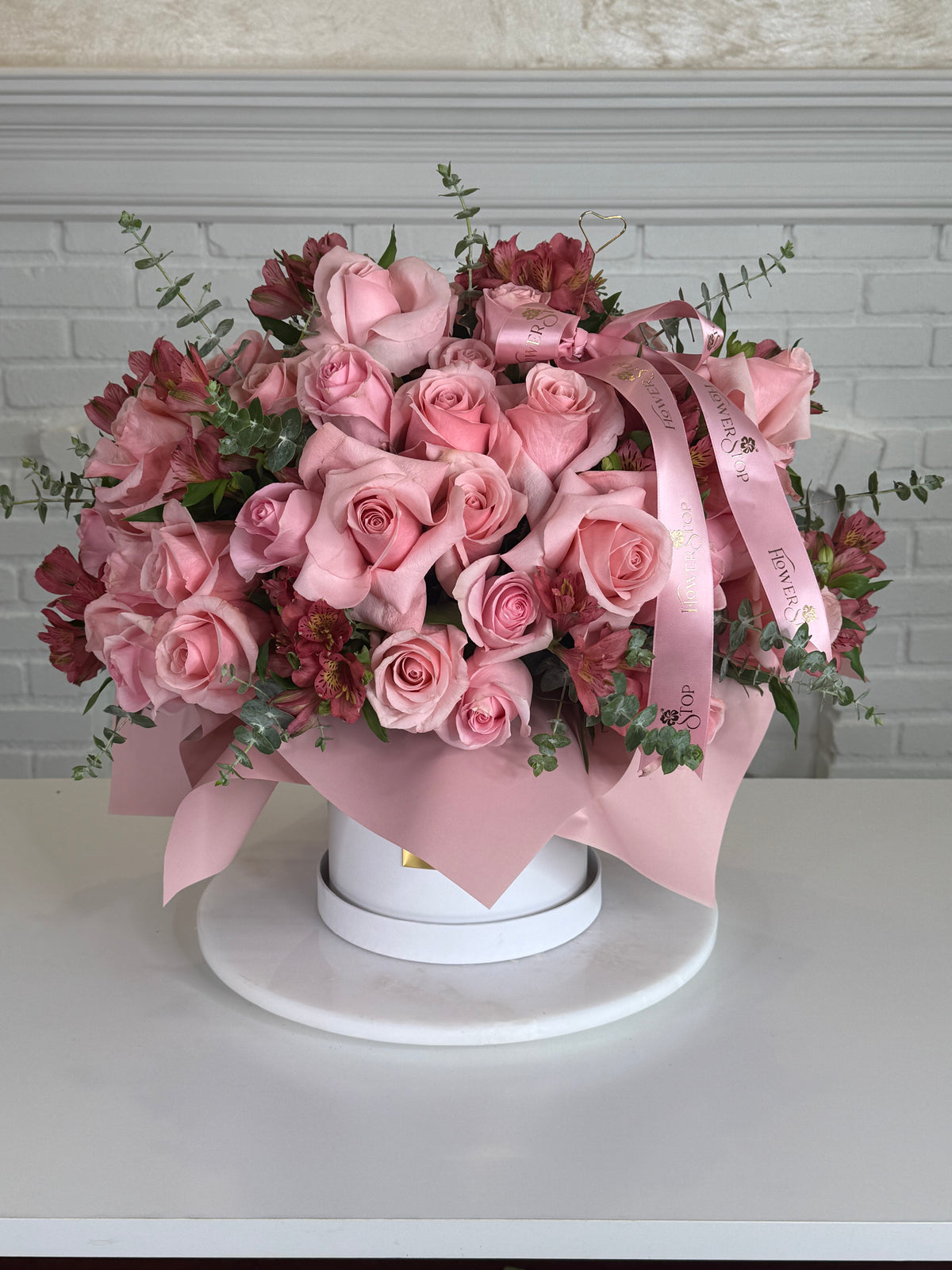 A floral arrangement of pink roses and burgundy alstroemeria with green foliage, wrapped in a light pink ribbon and presented in a round white box on a white pedestal against a white brick wall background.