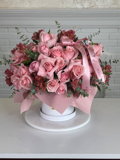 A floral arrangement of pink roses and burgundy alstroemeria with green foliage, wrapped in a light pink ribbon and presented in a round white box on a white pedestal against a white brick wall background.